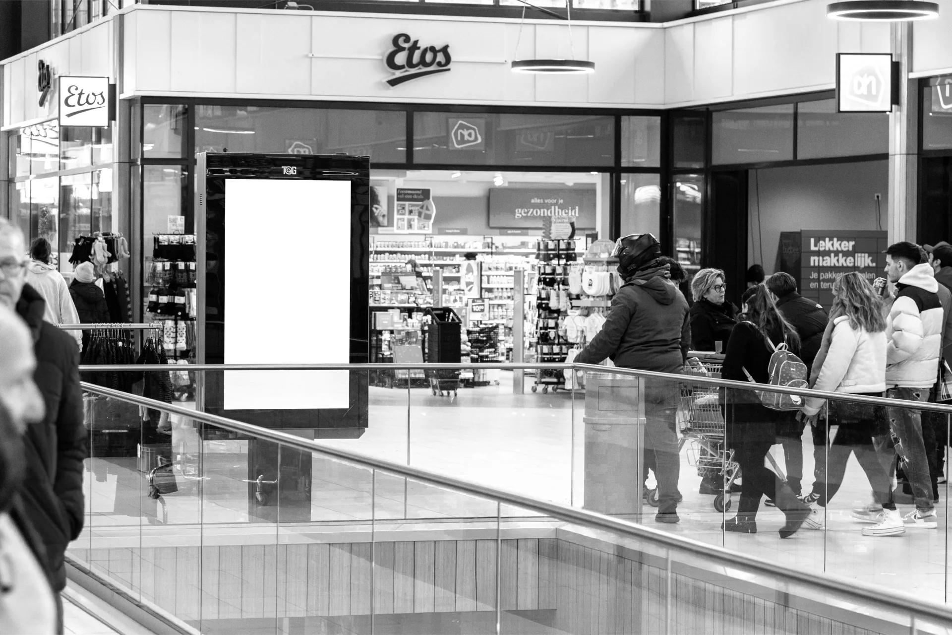 Shoppers passing a digital screen in a mall concourse, beside the shop fronts it points at