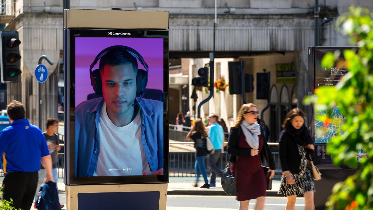 Un écran de rue diffusant une campagne de recrutement, devant lequel passent des navetteurs